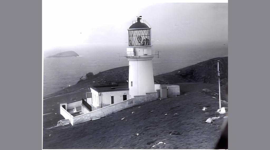 Black-and-white image of the Flannan Isles Lighthouse standing on a remote rocky island, surrounded by foggy sea and empty coastal cliffs.